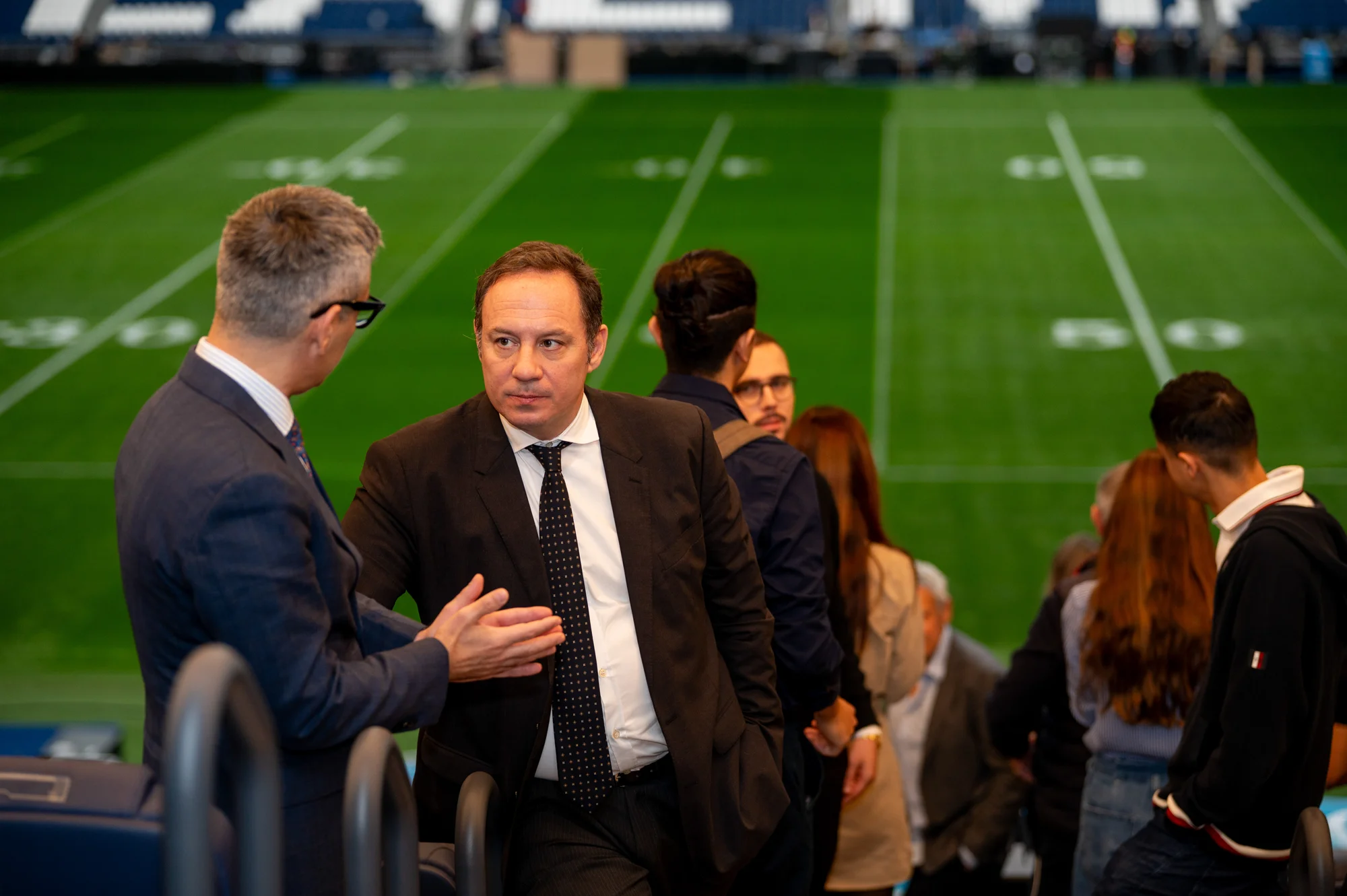 Invitados con traje conversando en la grada de un estadio durante un evento corporativo