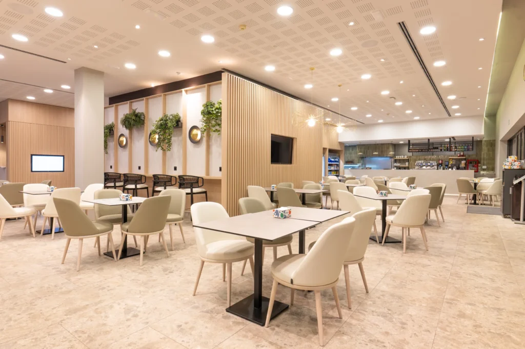 Cafetería luminosa de un club deportivo con mesas blancas y plantas colgantes