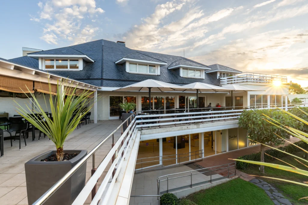 Edificio principal de un club deportivo con terraza y jardín bajo un cielo azul