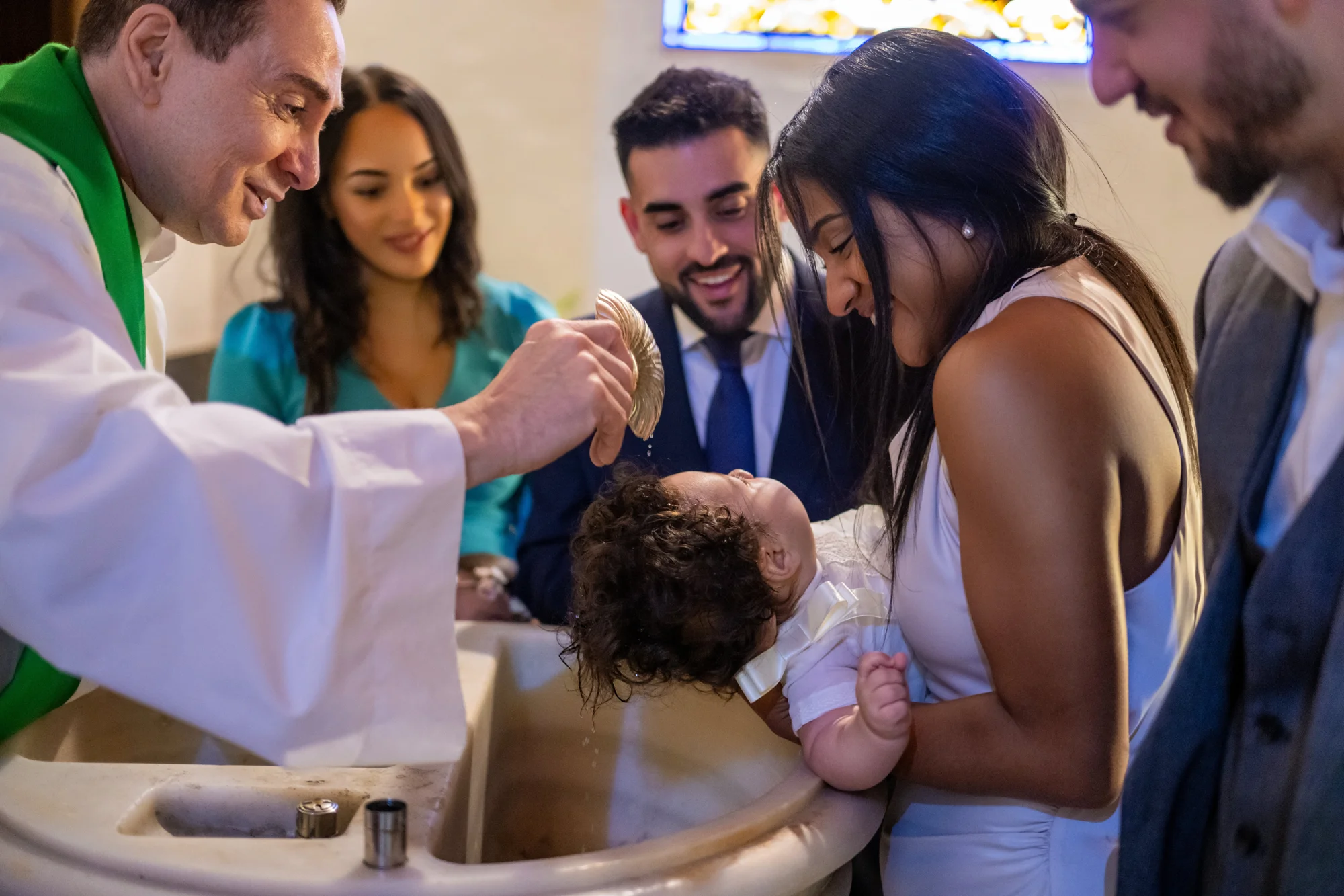 Sacerdote echando agua bautismal al bebé mientras la familia sonríe alrededor de la pila