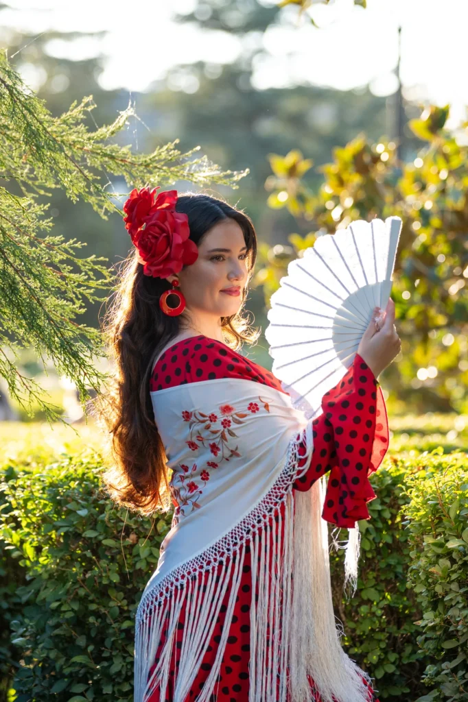 Mujer con flor roja en el pelo, mantón bordado y abanico blanco en un jardín de Madrid