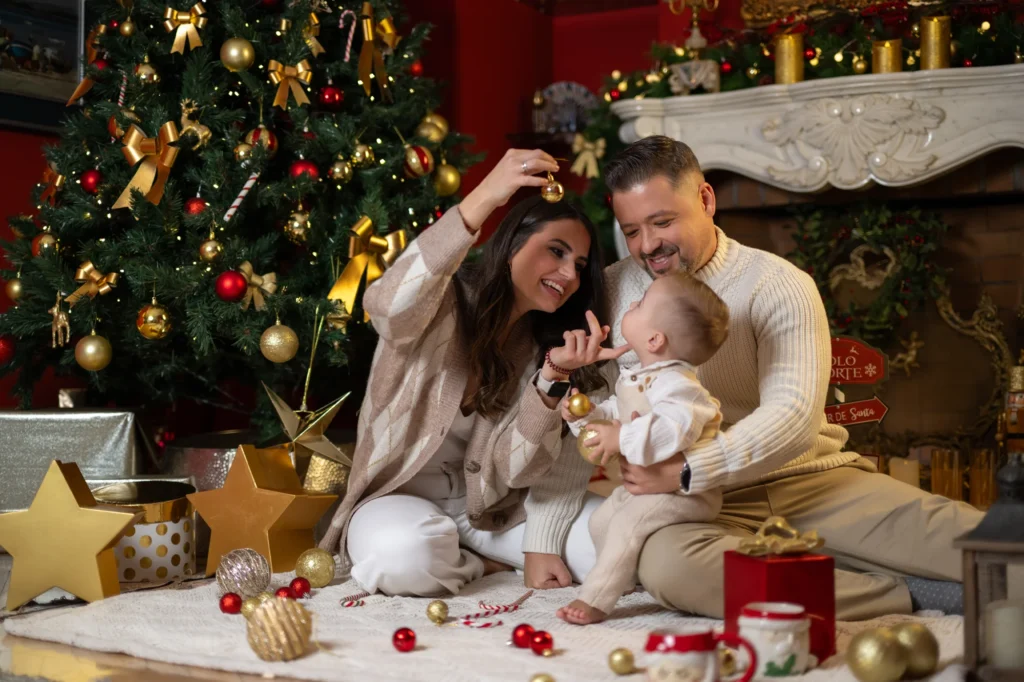 Familia sentada en el suelo jugando con su bebé junto al árbol de Navidad y la chimenea