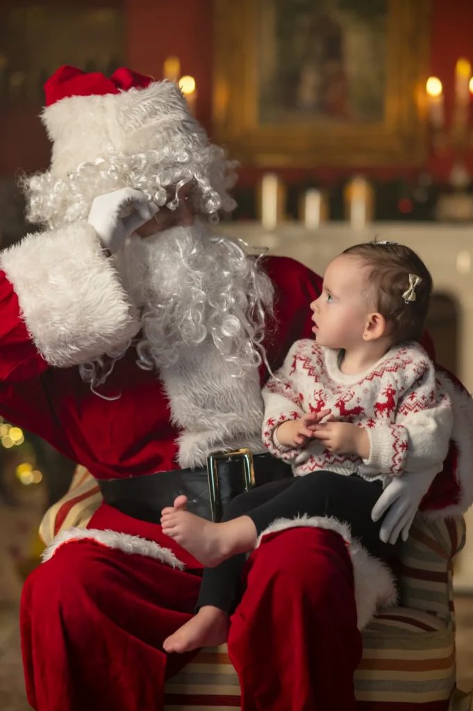 Niña pequeña sentada en el regazo de Papá Noel junto a la chimenea en una sesión de Navidad