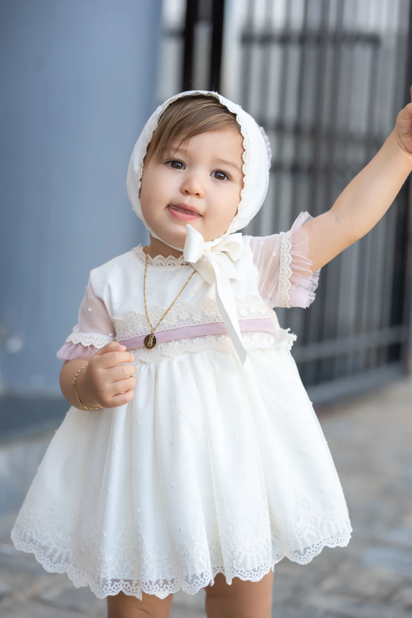 Niña pequeña con vestido y capota blancos saludando con la mano en su bautizo