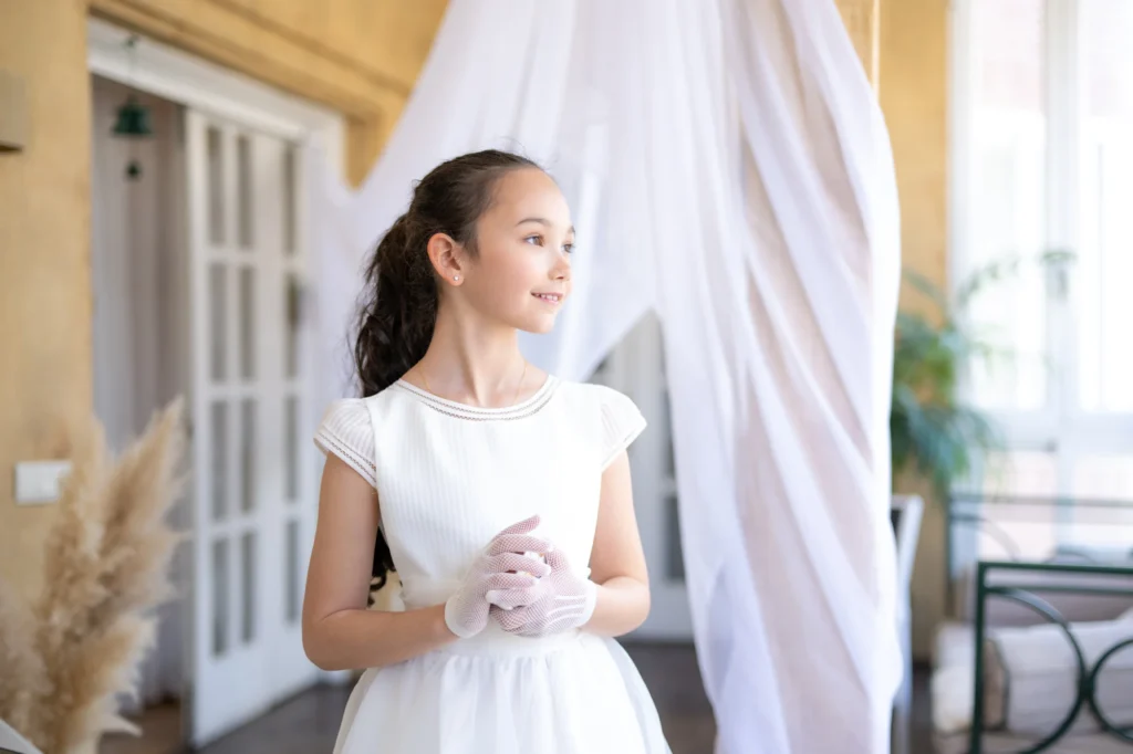 Niña con vestido de comunión blanco y guantes de encaje junto a cortinas blancas