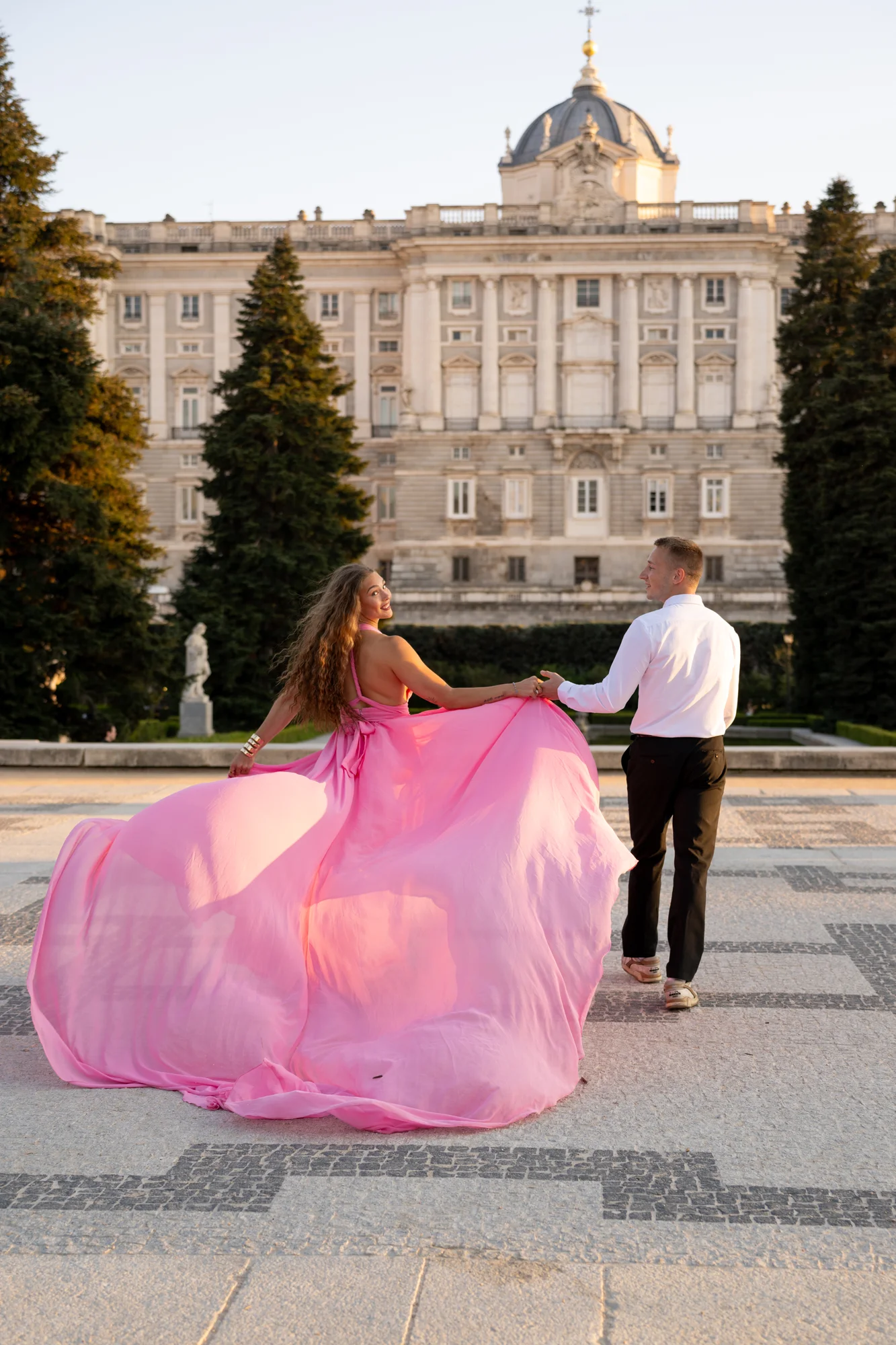 Pareja de la mano, ella con vestido rosa de cola larga, frente al Palacio Real de Madrid