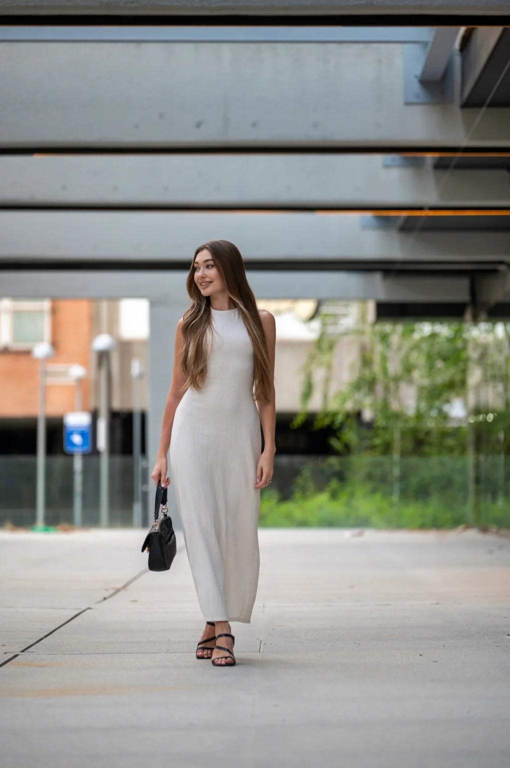 Modelo con vestido blanco largo y bolso negro caminando bajo una marquesina moderna