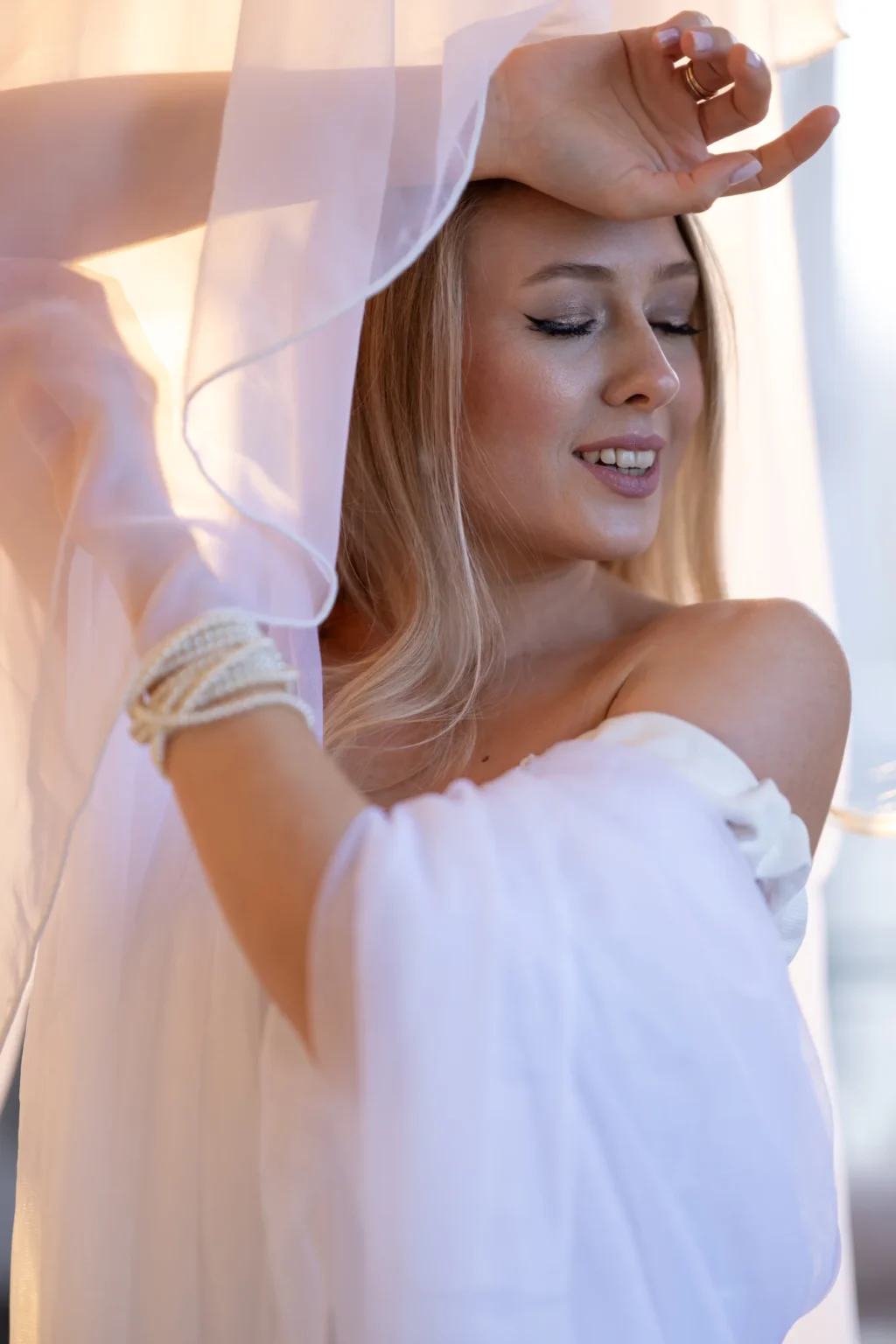 Modelo con vestido blanco y velo de gasa junto a una ventana luminosa