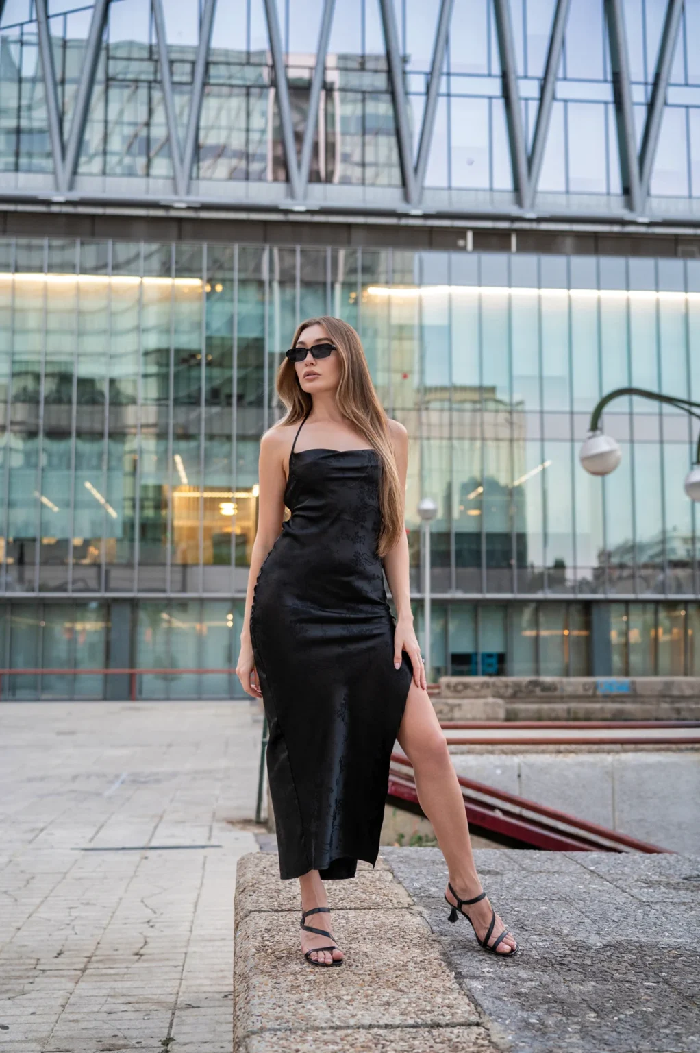 Modelo con vestido negro de abertura lateral posando ante un edificio de oficinas acristalado