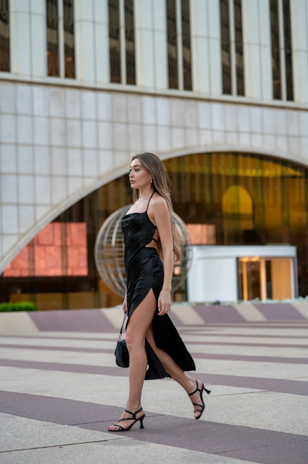 Modelo con vestido negro caminando por una plaza frente a un edificio con arco de cristal