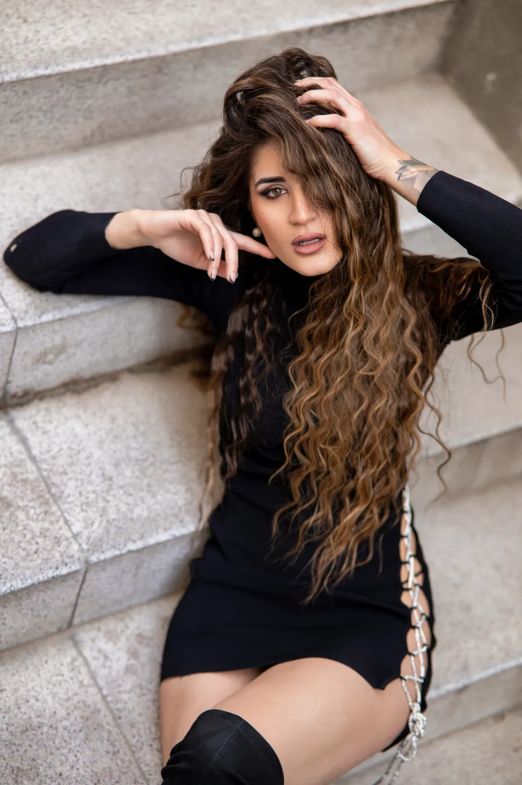 Modelo con vestido negro corto y melena rizada recostada en unas escaleras de piedra