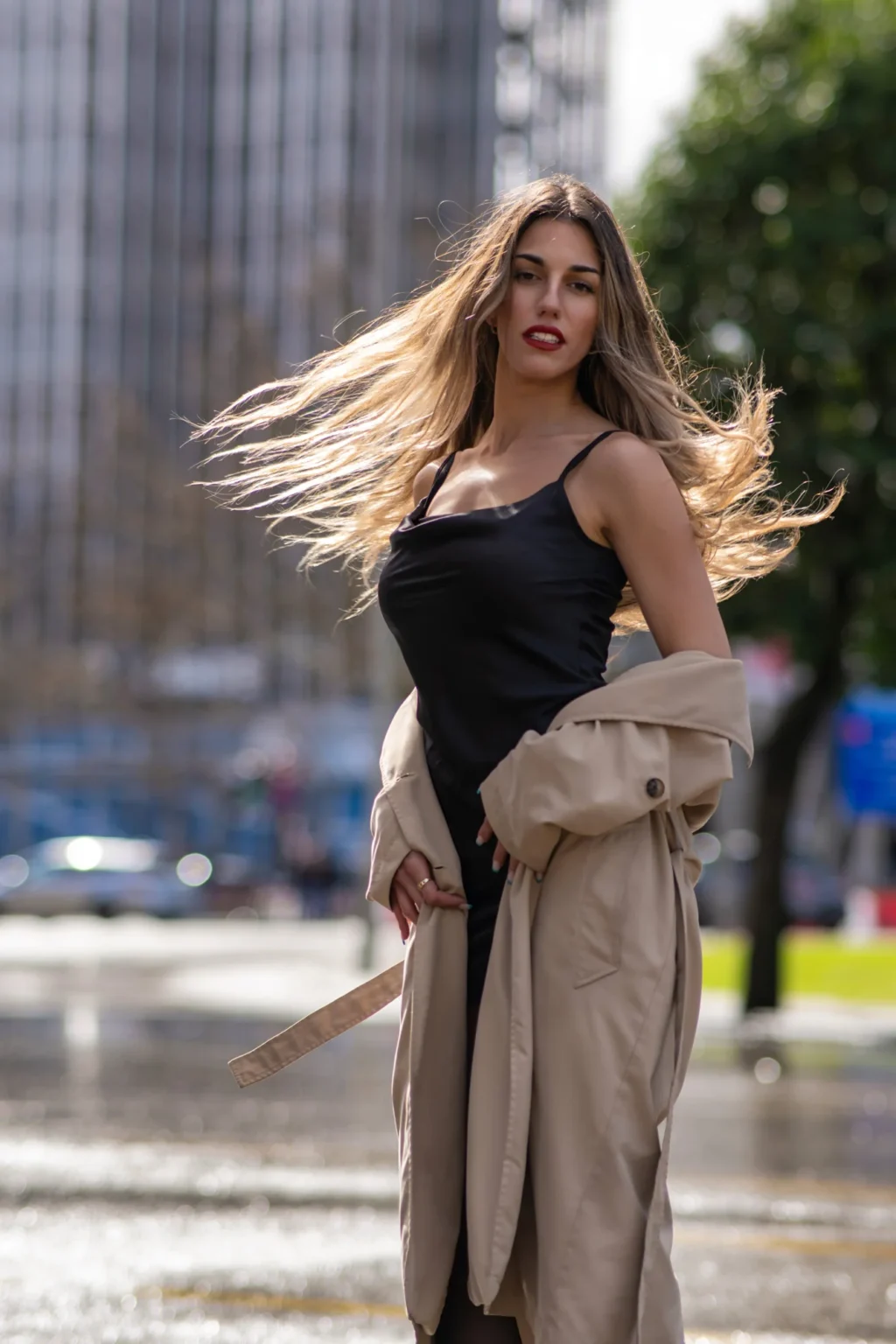 Modelo con vestido negro y gabardina con el pelo al viento en una calle de Madrid