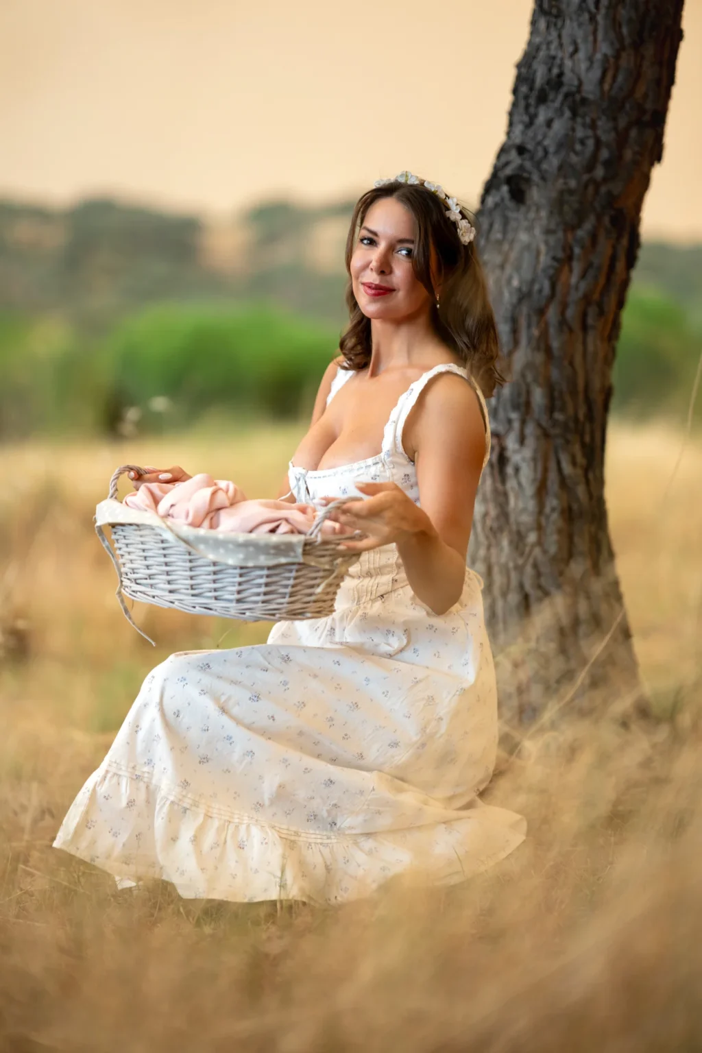 Mujer con vestido blanco y cesta de mimbre sentada bajo un árbol en un campo al atardecer