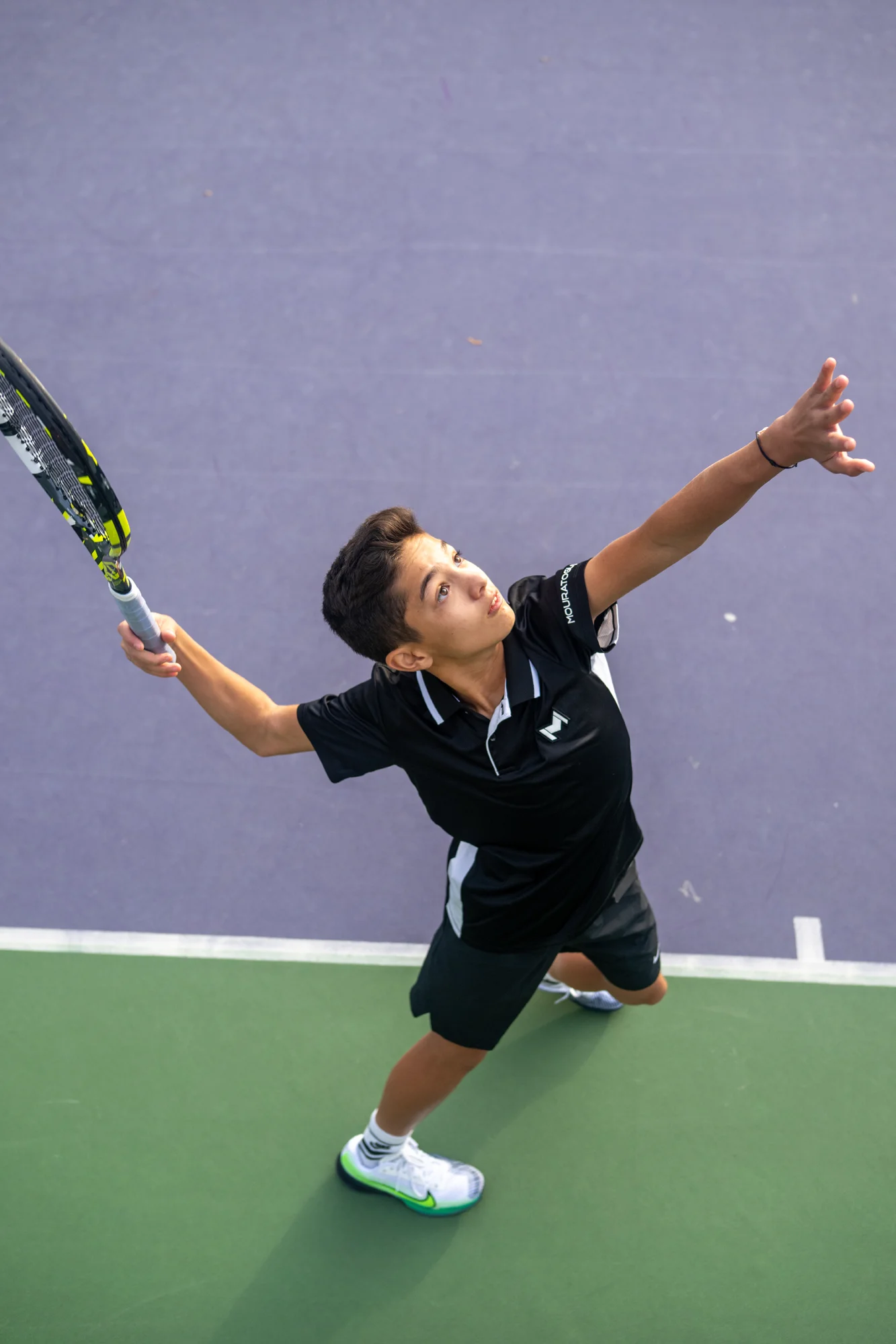 Niño golpeando la pelota de tenis en un saque sobre una pista verde y pared morada