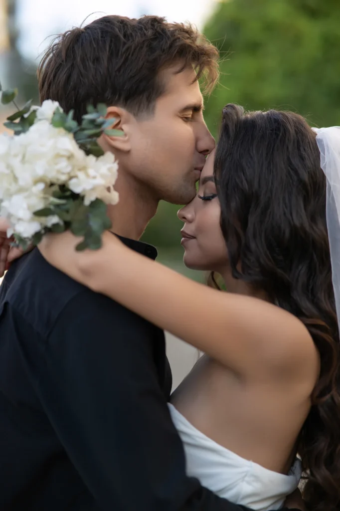 Novio besando la frente de la novia, ella con flores blancas en el pelo