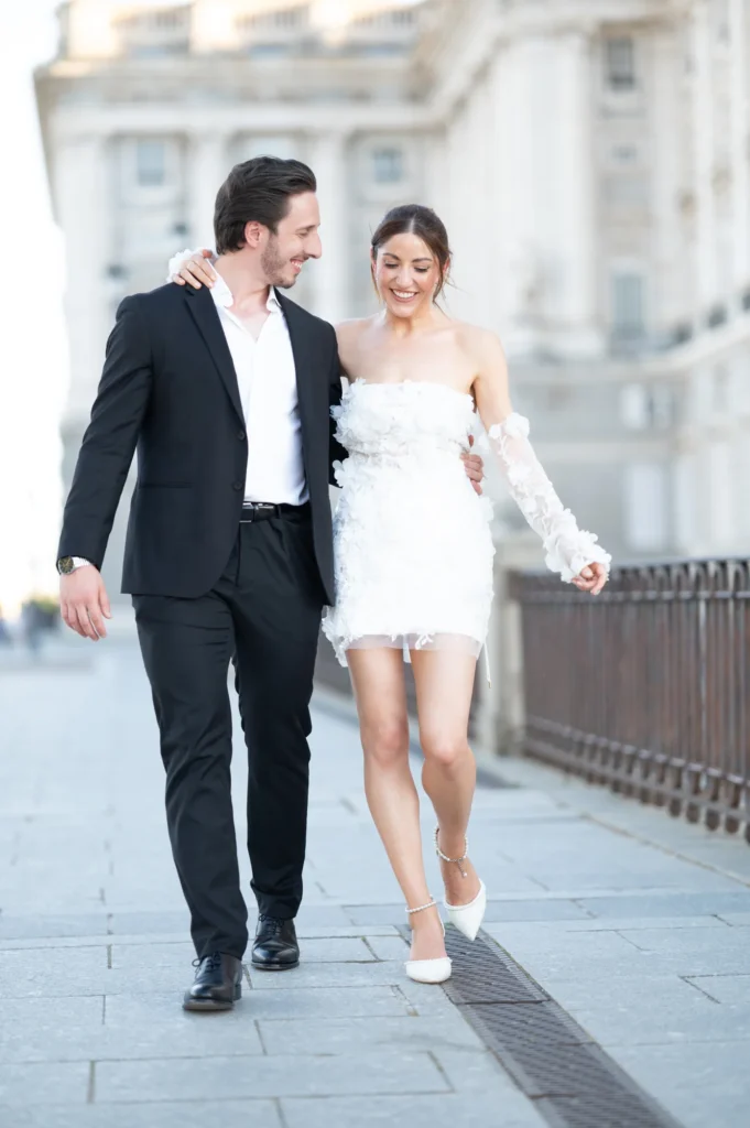 Pareja caminando de la mano junto a la barandilla del Palacio Real, ella con vestido corto blanco