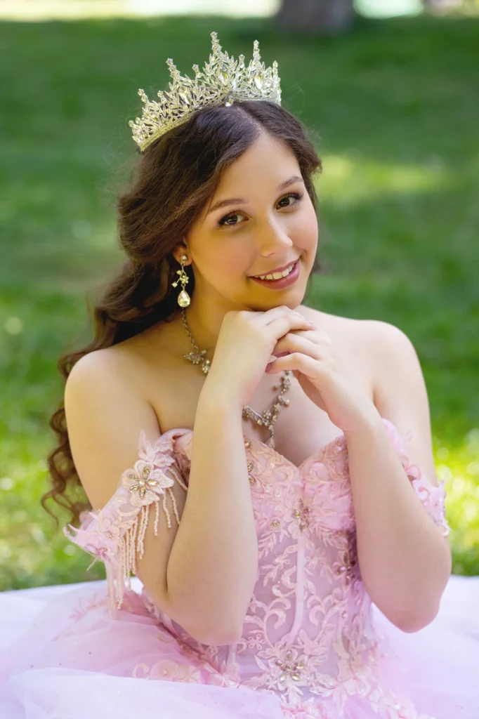 Quinceañera con corona apoyando la cara en las manos, con vestido rosa y fondo de jardín