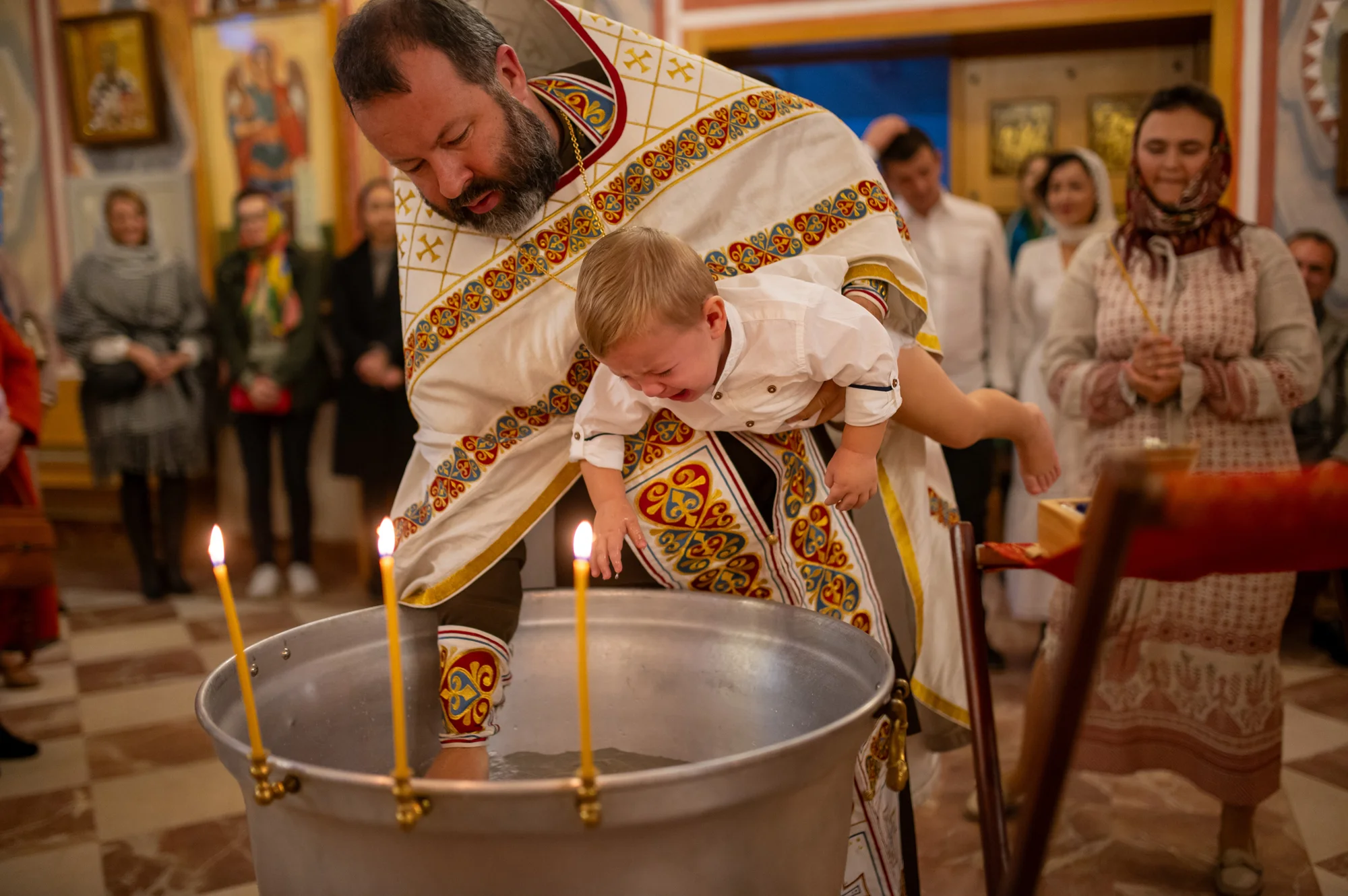 Sacerdote ortodoxo sumergiendo al bebé en la pila bautismal rodeado de velas