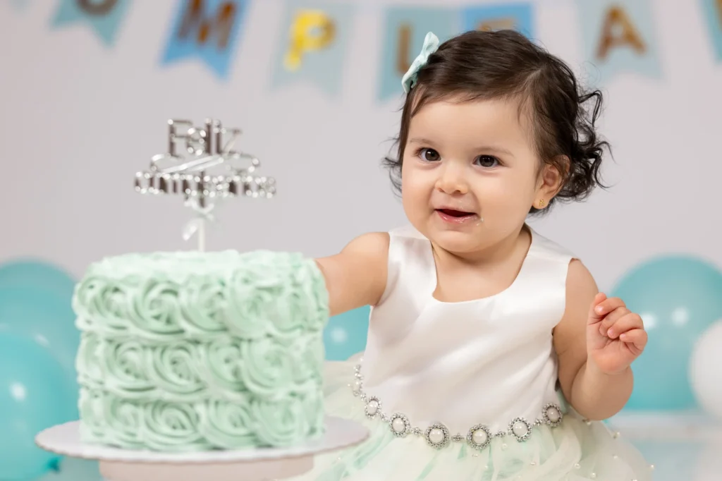 Bebé sonriente junto a su tarta de primer cumpleaños con globos turquesa de fondo