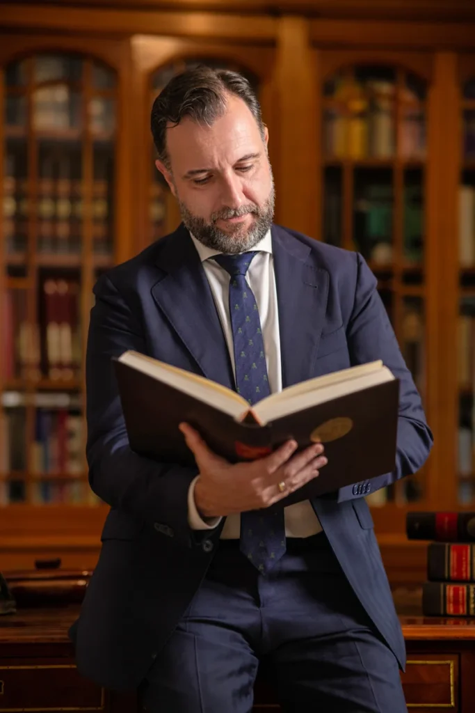 Hombre con traje leyendo un libro de pie en una biblioteca con estanterías de madera