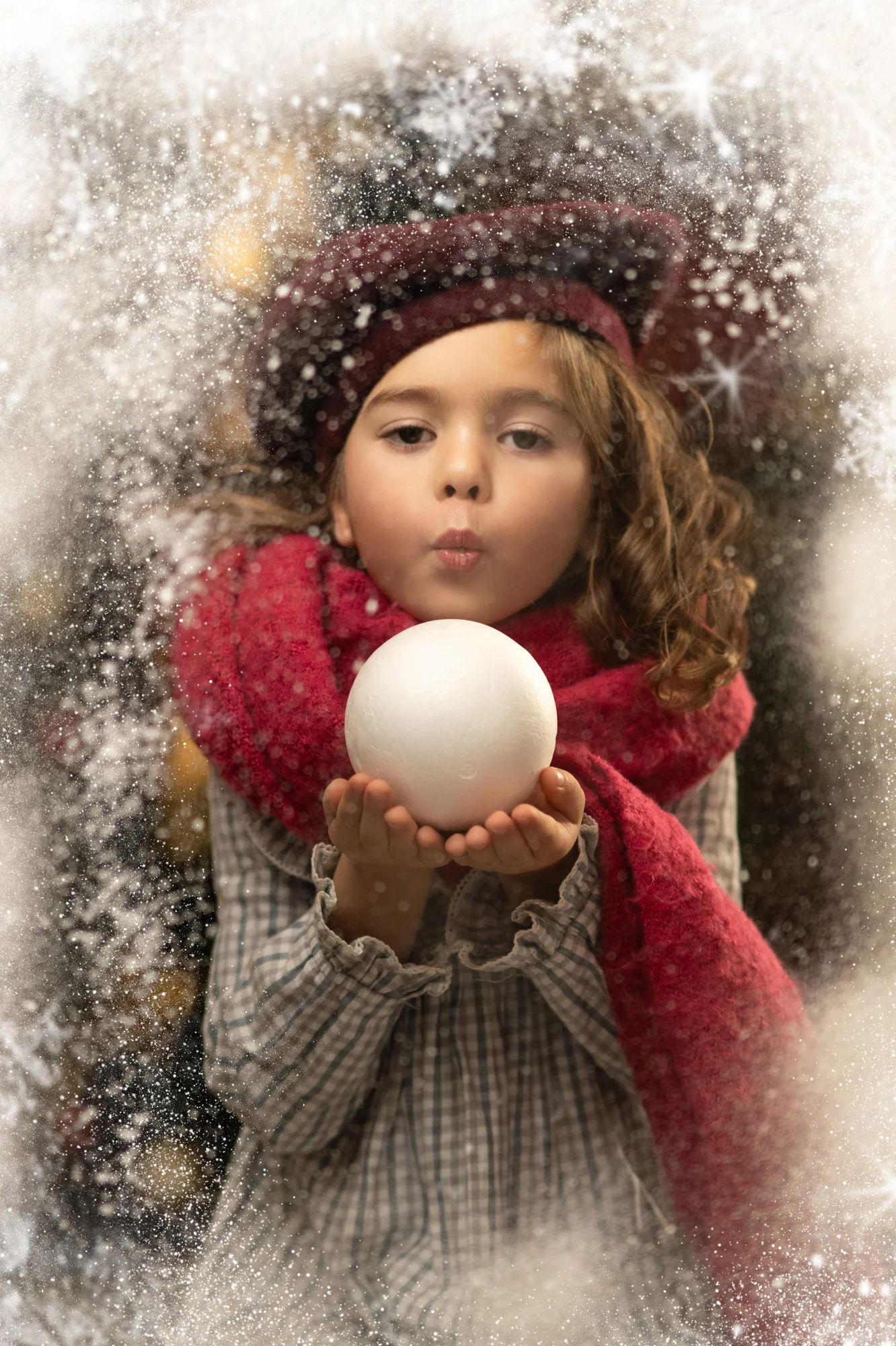 Niña con gorro y bufanda roja soplando una bola de nieve en una fotografía navideña