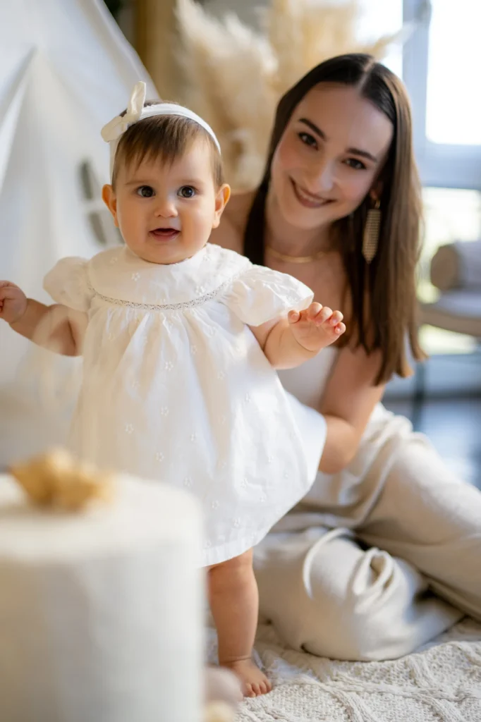 Bebé con vestido blanco dando sus primeros pasos de la mano de su madre en el estudio