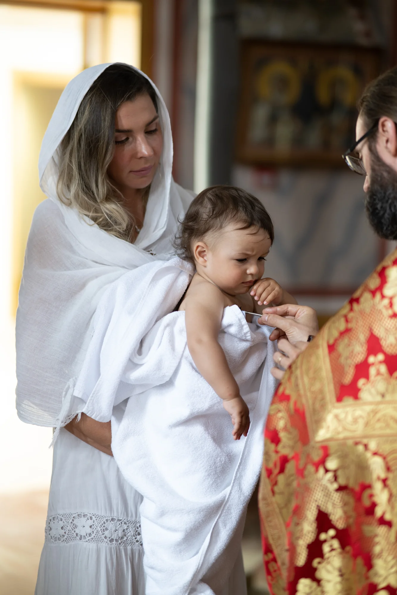Madre con velo sosteniendo al bebé envuelto en una toalla blanca junto al sacerdote