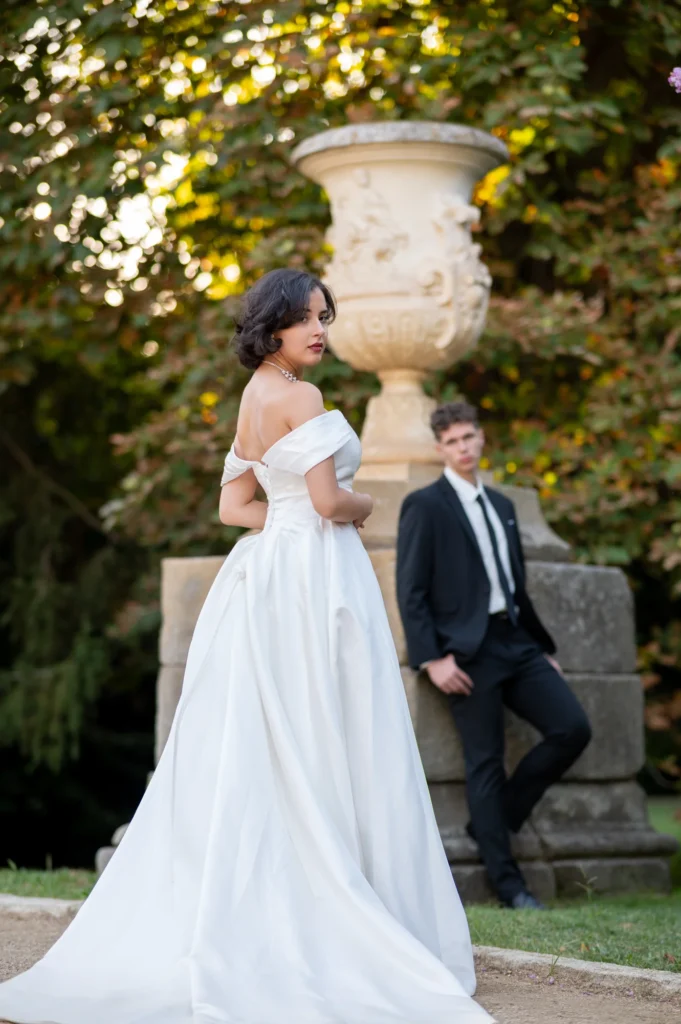 Novia con vestido blanco de cola y novio de traje junto a un jarrón de piedra en un jardín