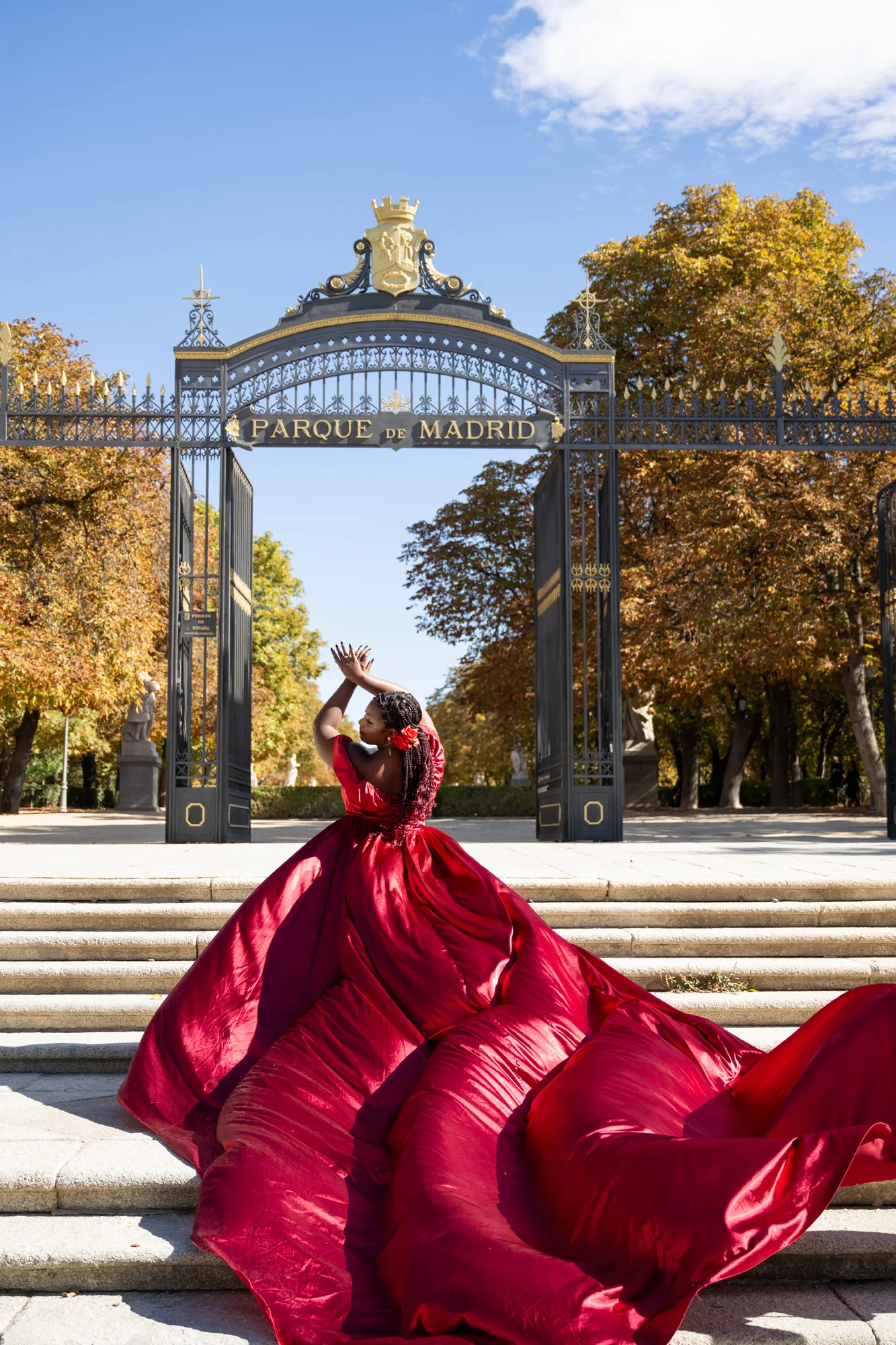 Mujer con vestido rojo extendido en el suelo ante una puerta de hierro del Parque del Retiro