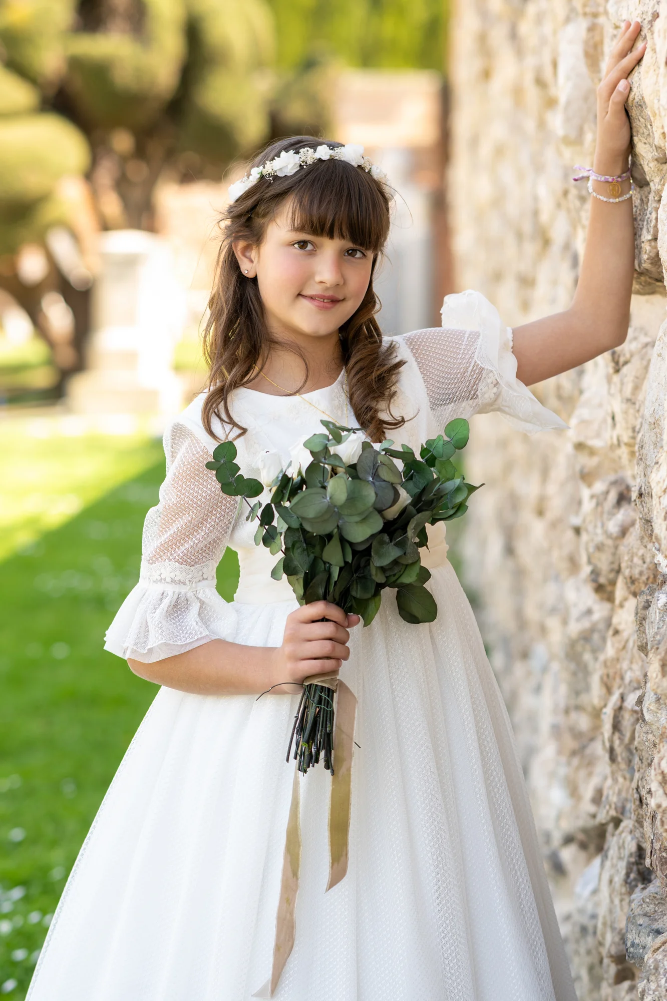 Niña con vestido de comunión y ramo de eucalipto apoyada en un muro de piedra en el jardín