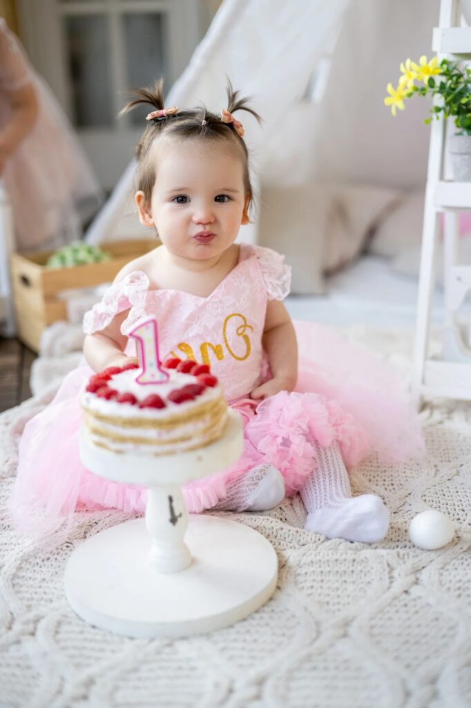 Bebé con coletas y tutú rosa junto a su tarta de cumpleaños con vela del número 1
