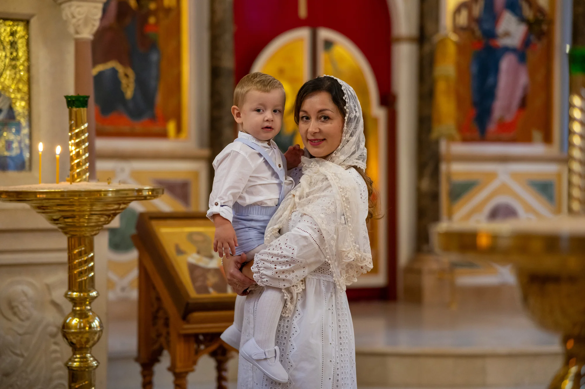 Madre con pañuelo blanco sosteniendo a su bebé frente al iconostasio de una iglesia ortodoxa